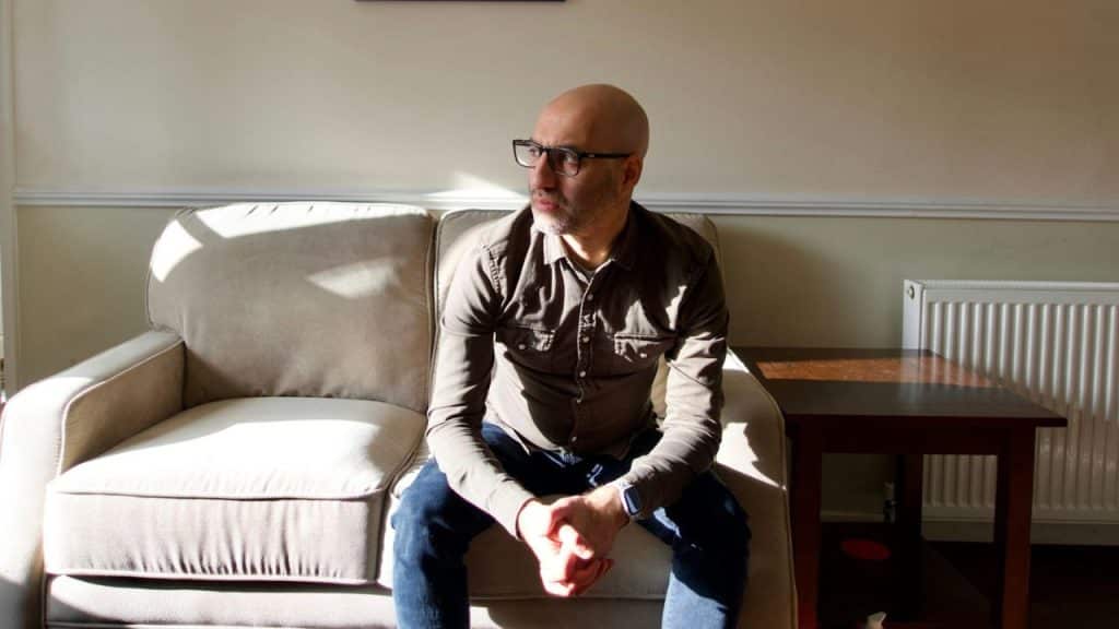 Bald man with a beard and glasses sitting on a couch, looking out a window with sunlight streaming in.