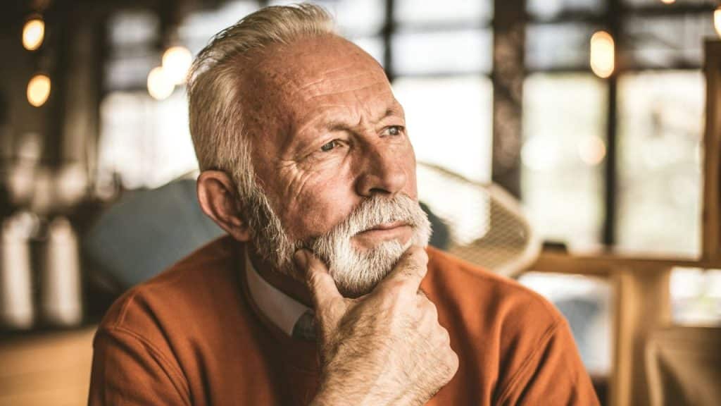 Older man with white beard in an orange sweater resting his chin on his hand.