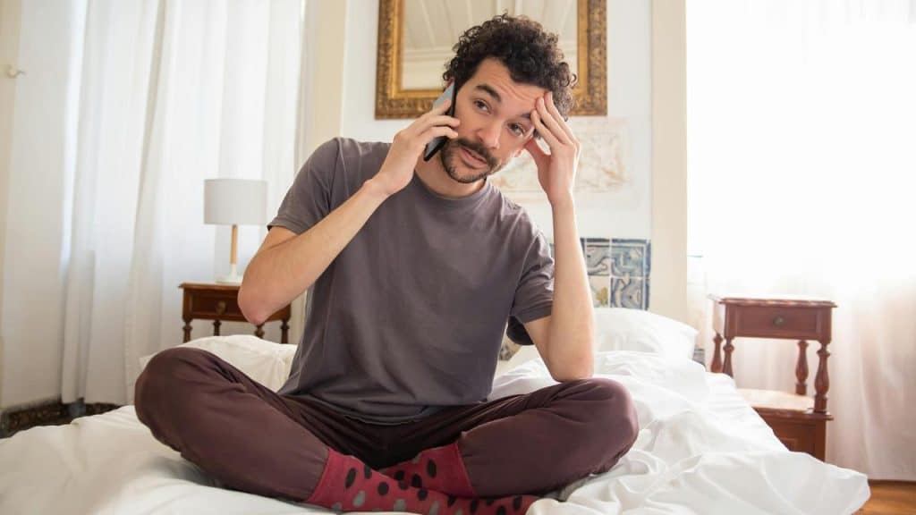 A man sitting cross-legged on a bed while talking on the phone and touching his forehead.
