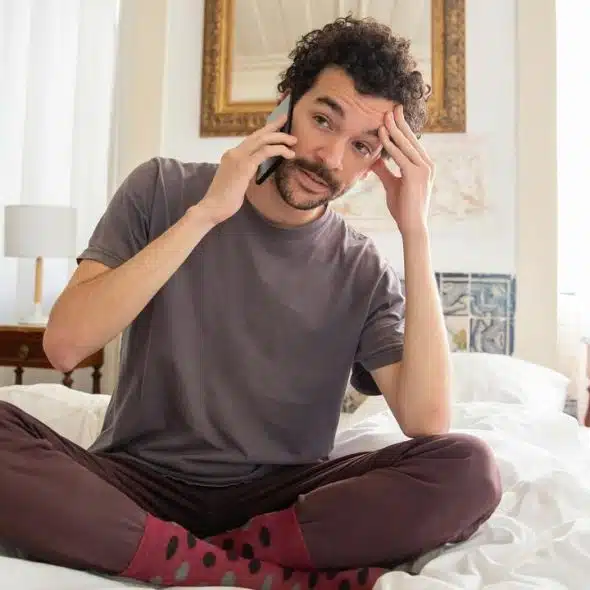 A man sitting cross-legged on a bed while talking on the phone and touching his forehead.