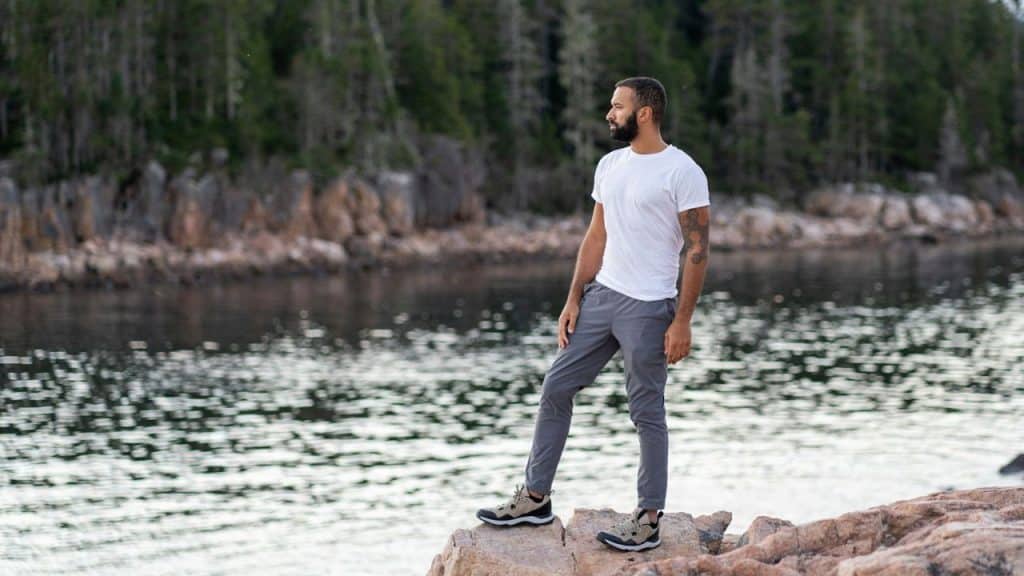 A fit man in a white shirt and gray pants stands on rocks by a lake.