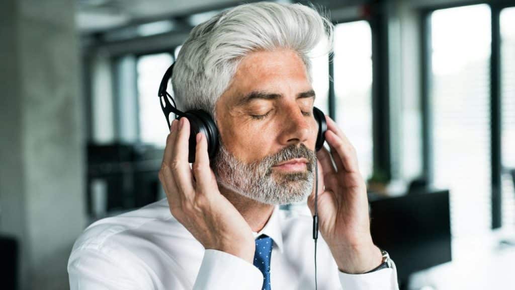 Older man in a white shirt and tie with his eyes closed, wearing over-ear headphones and holding them with both hands.