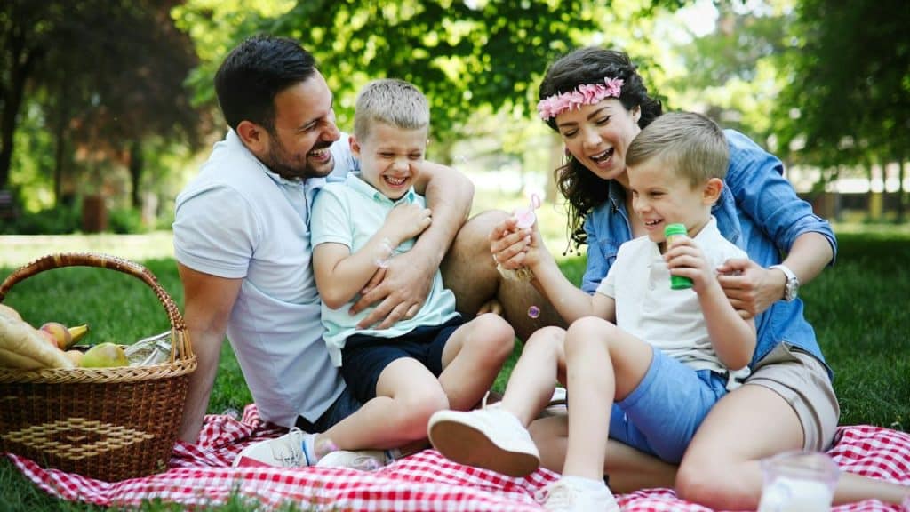 Family of four laughing on a red checkered blanket during an outdoor picnic.