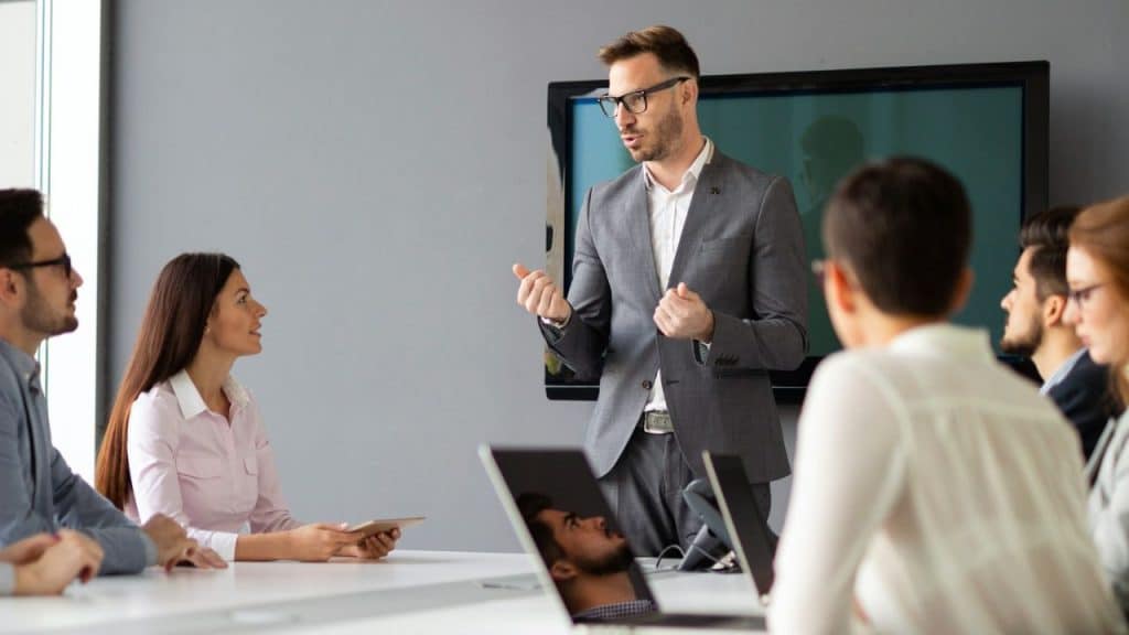 Man in a gray suit and glasses presenting to a group of people in a meeting room.