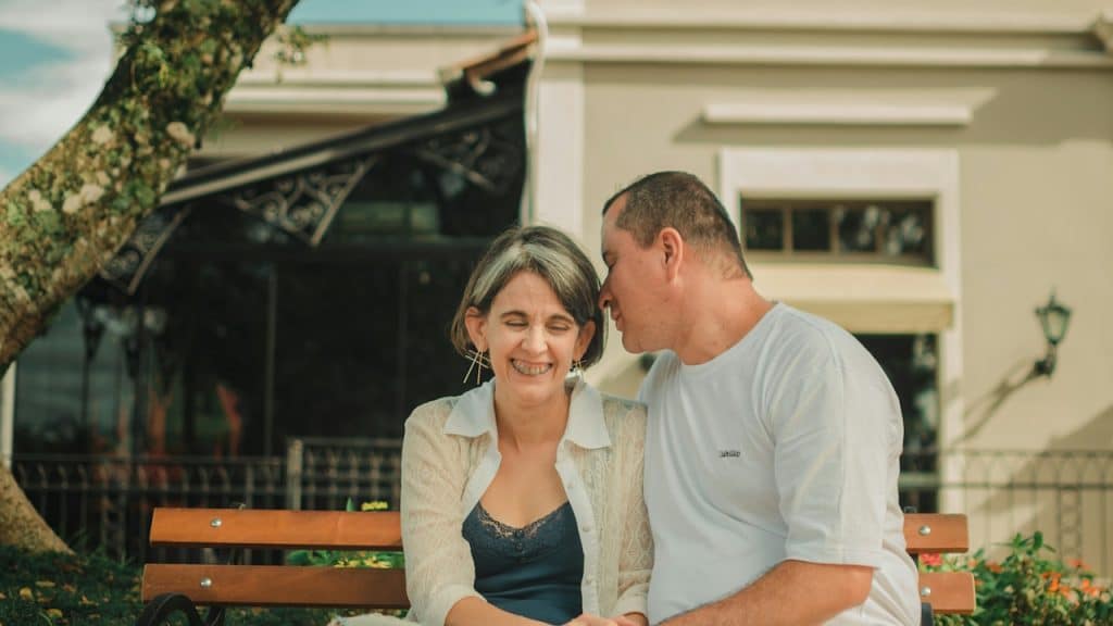A couple sitting close together on an outdoor bench, holding hands as the man whispers to the woman who is laughing warmly.