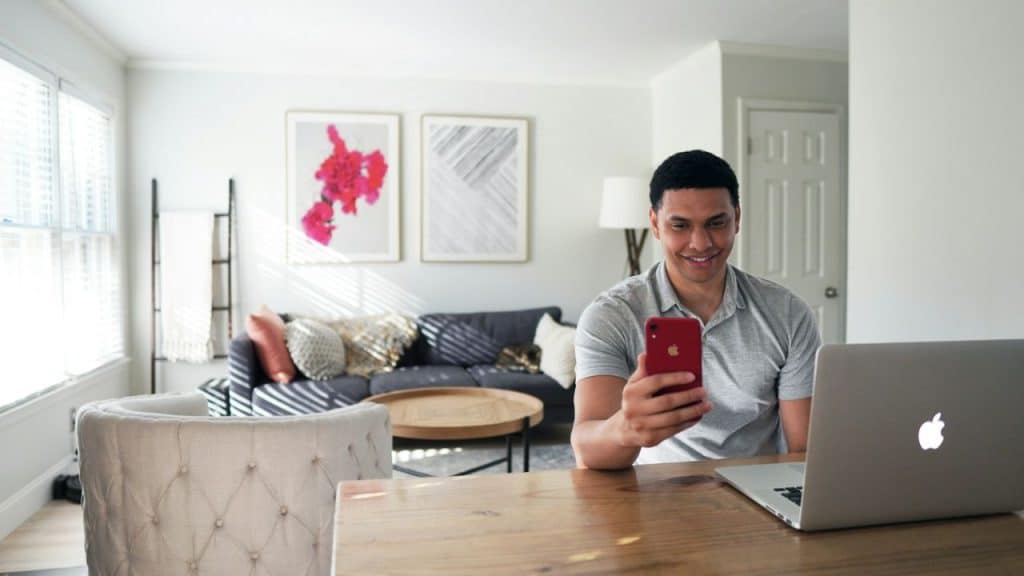 A smiling man sits at a wooden table in a bright living room, holding a red smartphone near his laptop.
