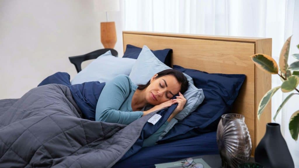 A woman with dark hair is sleeping on her side under a dark blue and grey blanket in a bed.