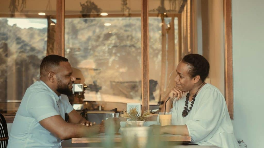 A man and woman are seated at a table in a bright cafe, smiling at each other during a conversation.