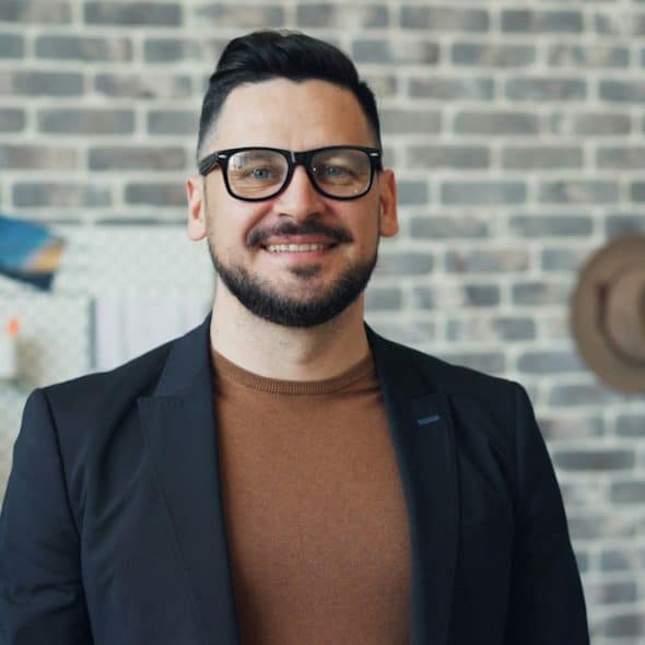A man wearing glasses standing in front of a brick wall.