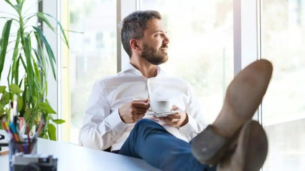 A bearded man in a white shirt has his feet up on a desk, holding a coffee cup, looking out a window.
