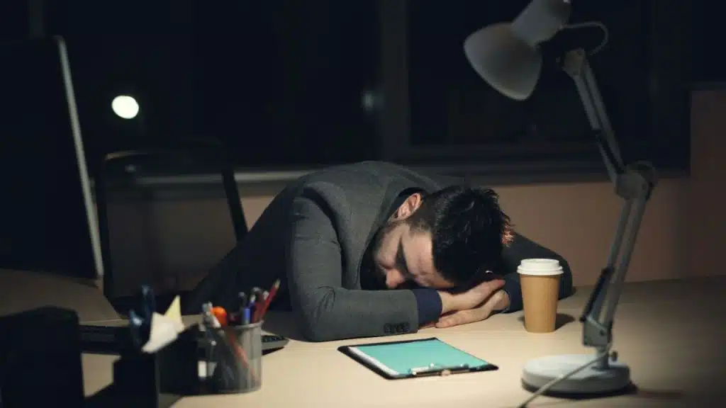 A man sleeping face down on a desk at night under a lamp, next to a coffee cup.