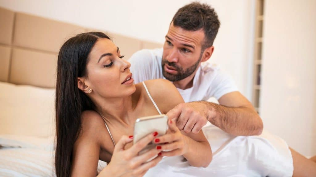 A serious man in a white t-shirt looks over a woman's shoulder as she uses her smartphone in bed.