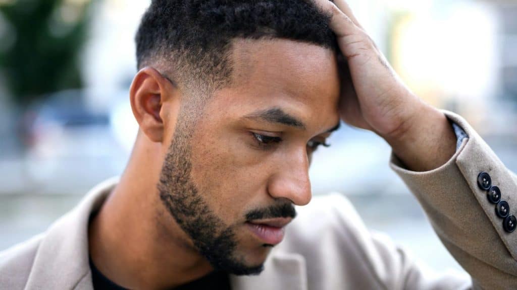 Close-up of a man in a beige jacket holding his hand on his head.