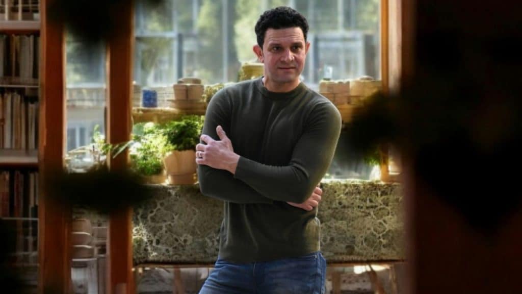 Man in a dark sweater and jeans standing indoors with his arms crossed.