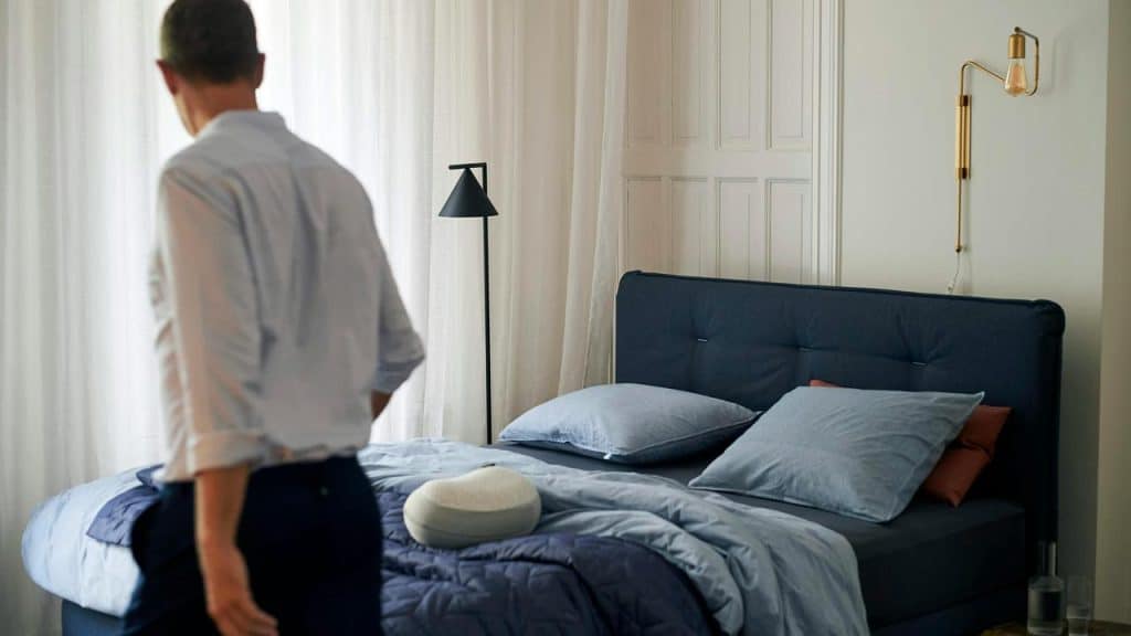 A man standing near a neatly made bed in a softly lit bedroom.