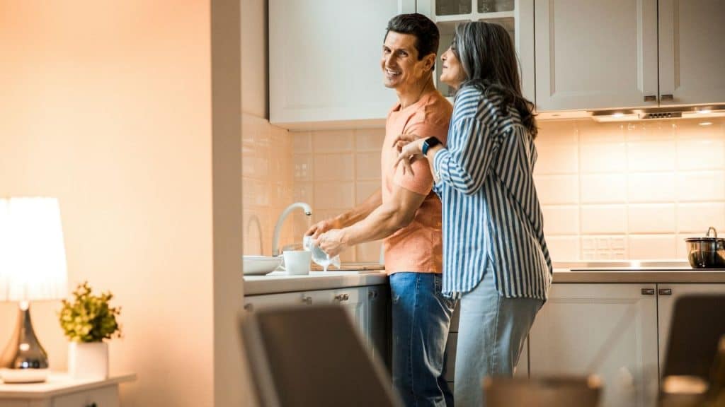 A smiling man is washing dishes at a sink in a kitchen while a woman with gray hair is talking.