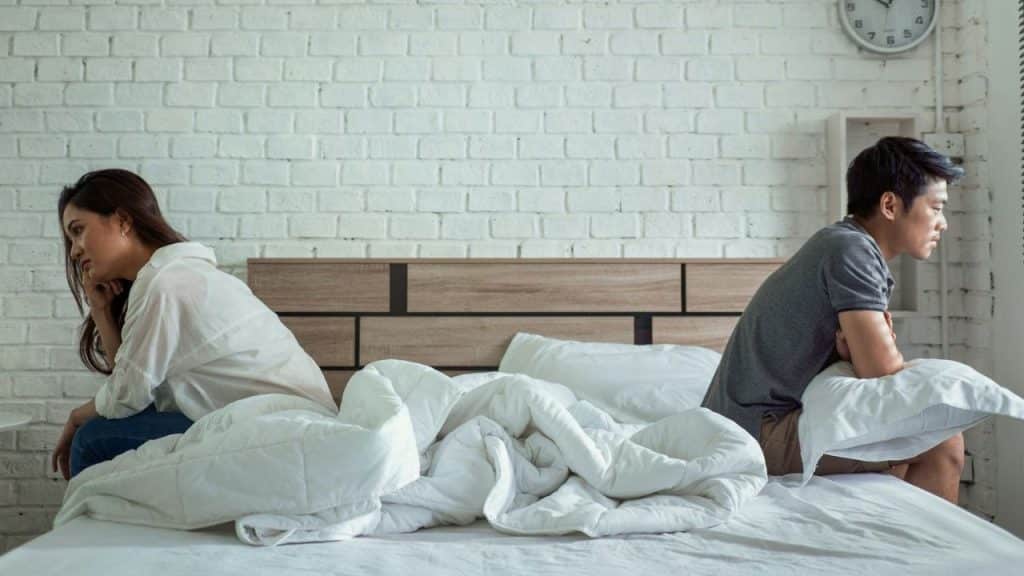 A man and a woman sit back-to-back on a bed, looking away from each other.
