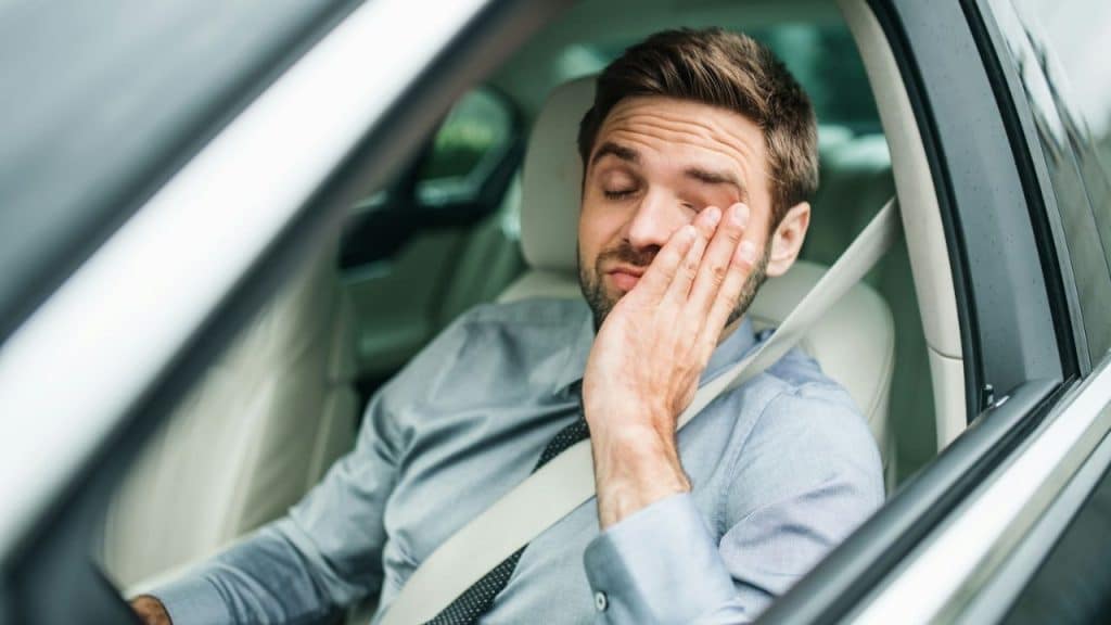 Man in a car, wearing a tie and seatbelt, rubs his closed eyes with his hand.