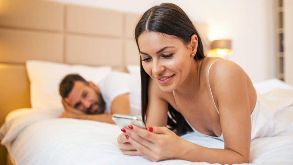 Woman smiling at her phone while a man lies in the background on the bed.