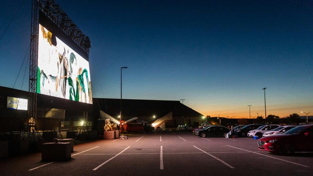 A drive-in movie theater with cars parked facing a large outdoor screen at sunset.
