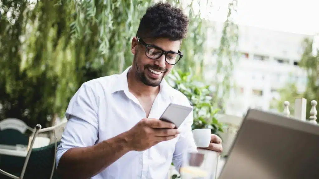 A man smiling while looking at his phone and holding a cup of coffee outdoors.