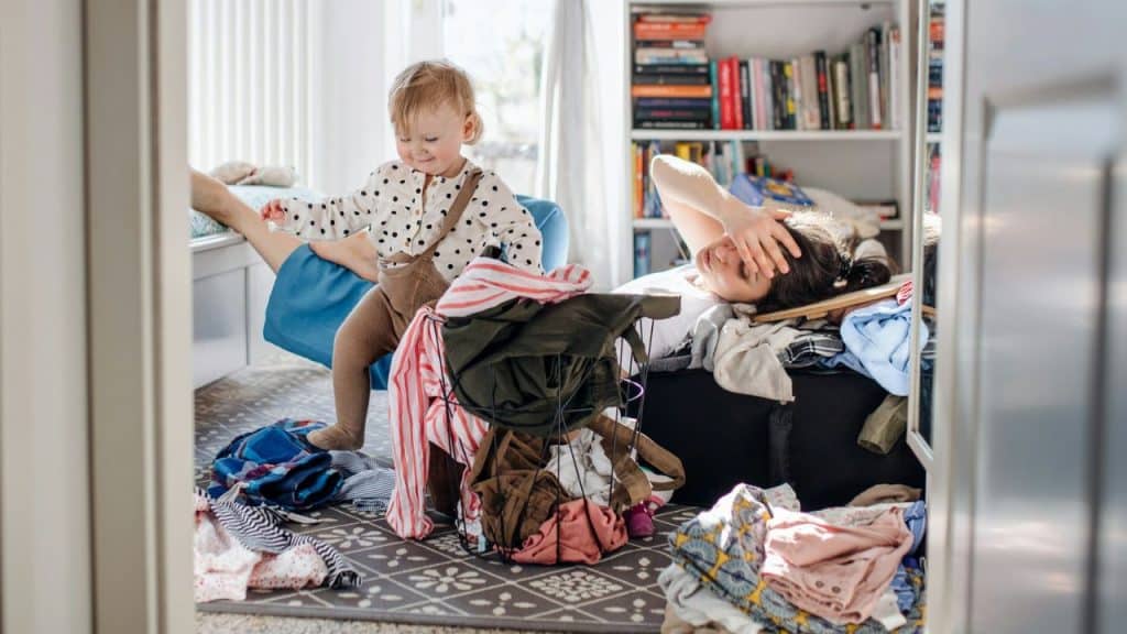 A small child plays among scattered clothes in a messy room while a woman lies down, covering her eyes.