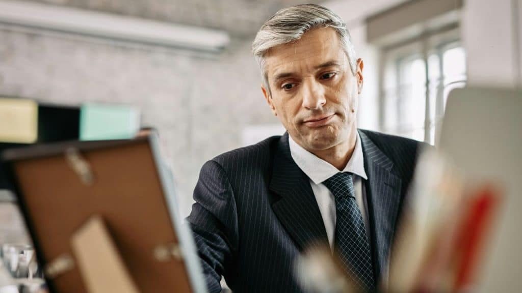 A middle-aged man in a pinstripe suit and tie looks down at a framed item on his desk.