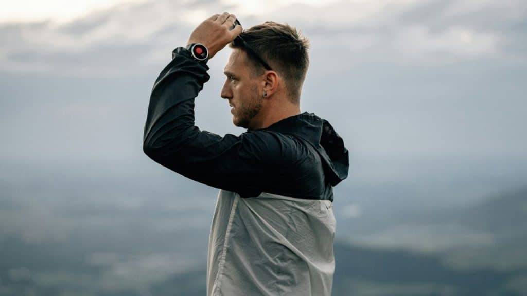 Man in a jacket and watch, looking into the distance with a cloudy sky backdrop.