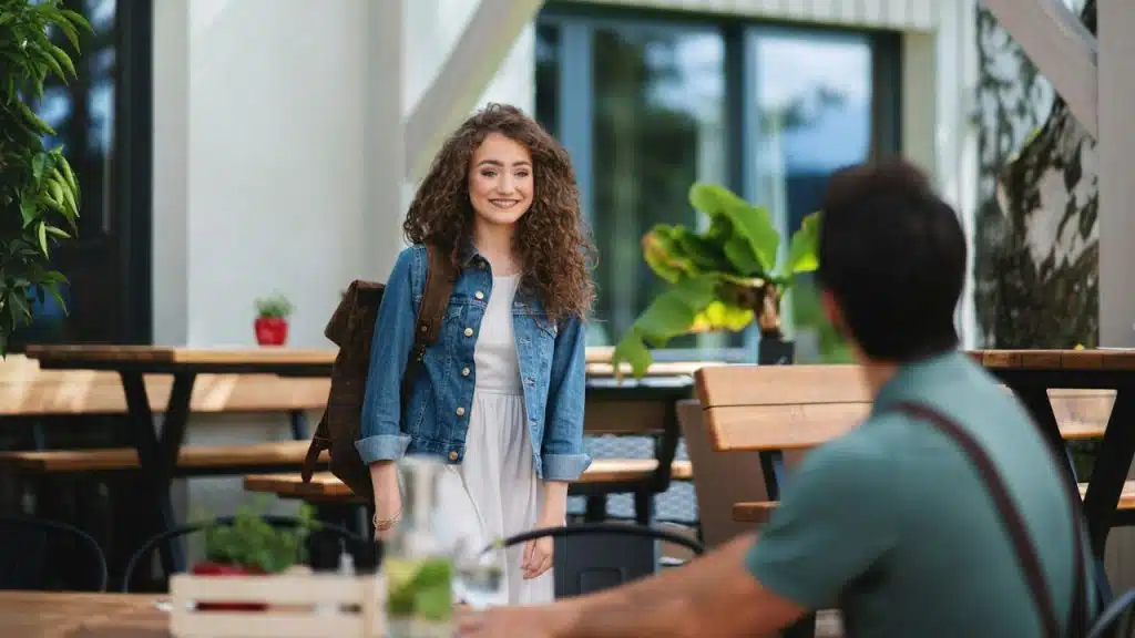 Smiling woman in denim jacket and backpack arriving at an outdoor cafe table.