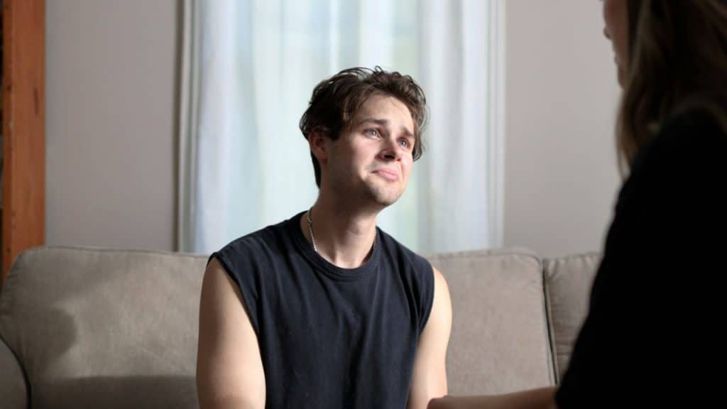 Young man sitting on a couch, looking distressed with watery eyes, facing a blurry figure.