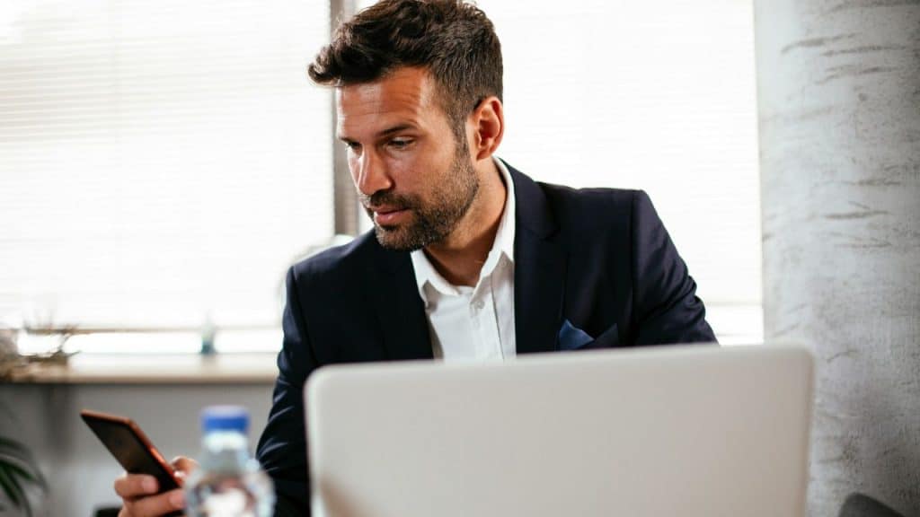 Man in a dark suit looking at a smartphone while sitting at a desk.
