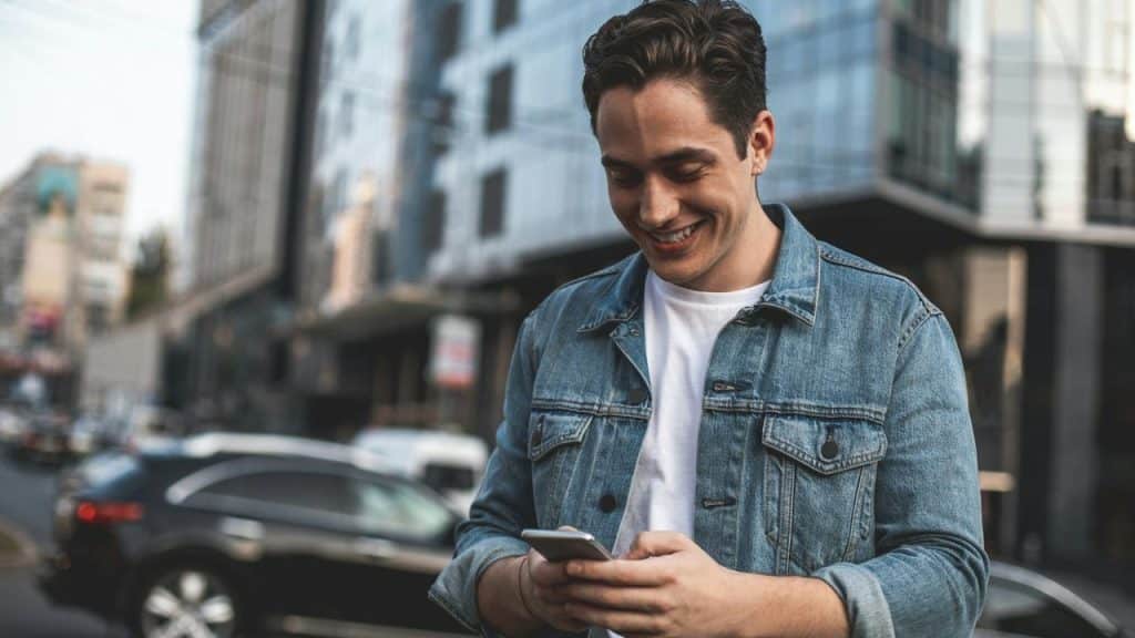 A man in a denim jacket smiling while looking at his phone on a city street.