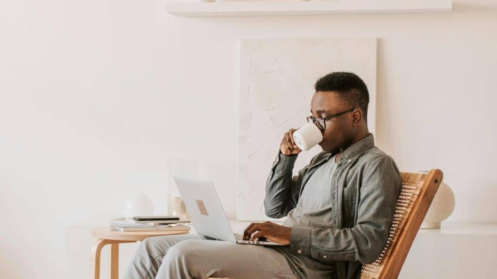 A person sitting on a chair, drinking from a cup while working on a laptop.