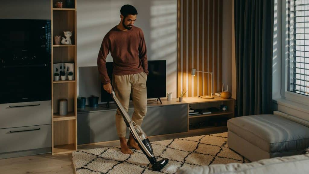 A man in a reddish-brown sweater and khaki pants is vacuuming a white, patterned area rug in a modern living room.
