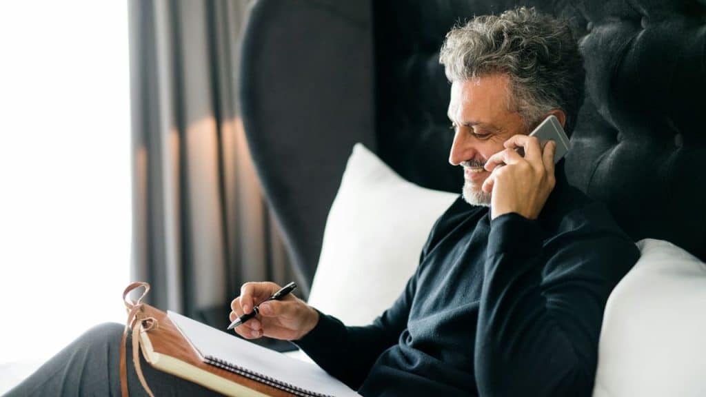 Mature man on a phone call, writing in a notebook while sitting in bed.