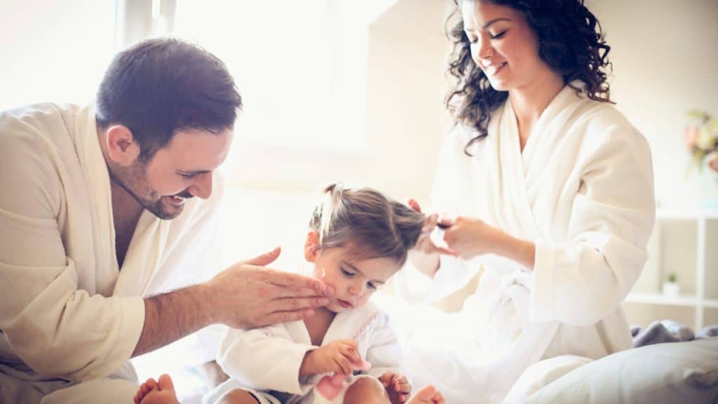 A young family in white bathrobes sits on a bed, with the mother styling the toddler's hair and the father gently touching the child's cheek.