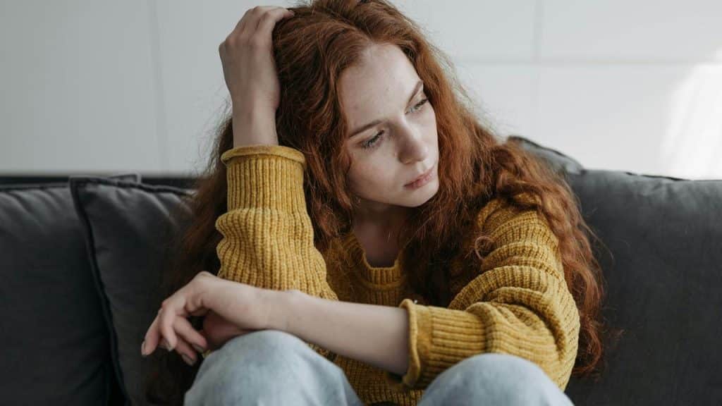 A young woman sitting on a couch looking thoughtful and resting her head on her hand.