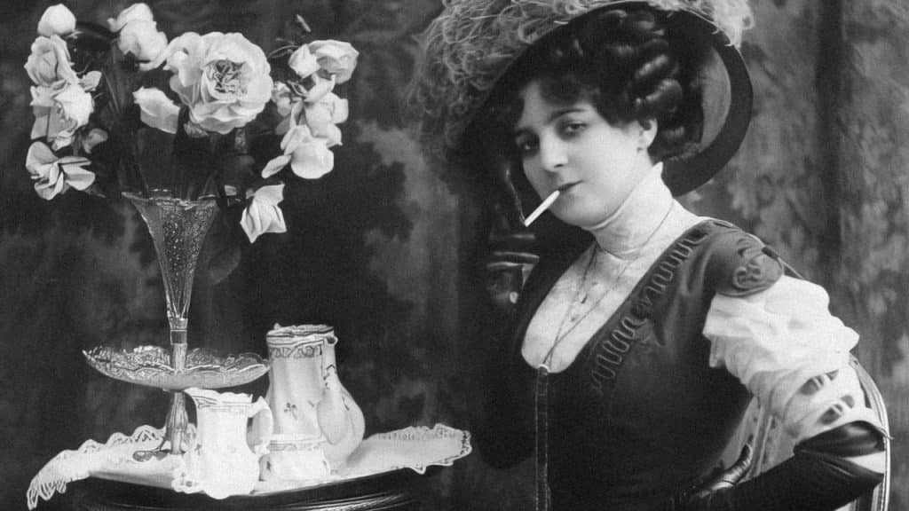 A black and white photo of a woman in a large hat with a cigarette in her mouth, next to a tray with a flower vase and creamer.