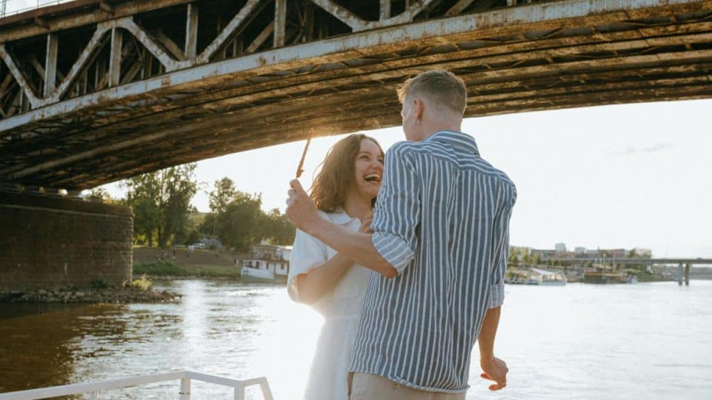 A smiling couple standing by the water under a bridge, holding hands.