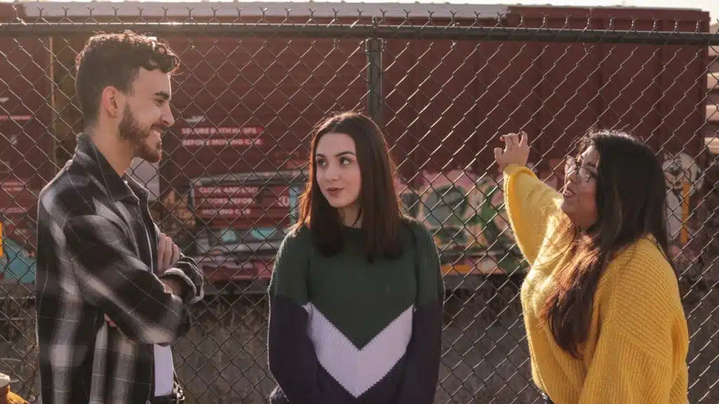 A group of three young adults talking outdoors in front of a chain-link fence and graffiti.
