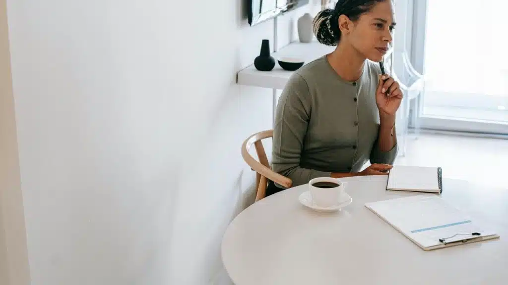 A woman sitting at a table with a cup of coffee holding a pen and looking thoughtful.