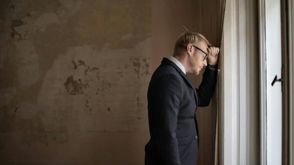 A man in a suit leaning against a window with his head lowered.