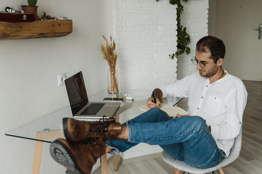 A Man Writing on a Notebook with Feet on Top of Desk