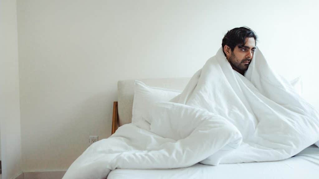 A man sitting in his bed while covered with a blanket and a sad expression on his face.