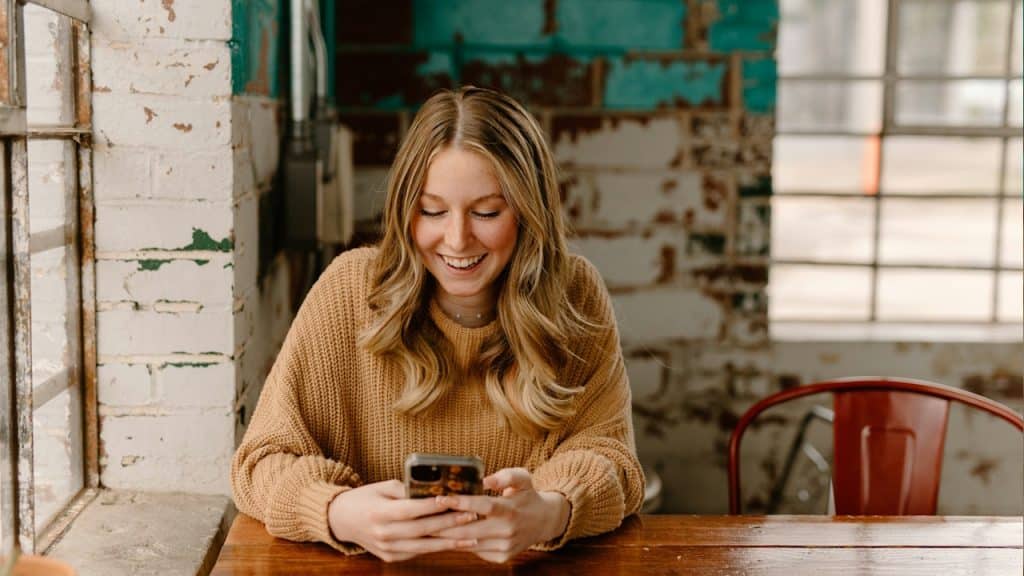 A smiling young woman with long blonde hair sits at a wooden table and looks down at her smartphone.