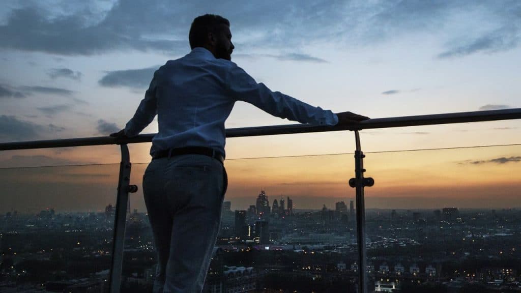 A man leans on a railing, silhouetted against a city skyline at sunset or twilight.