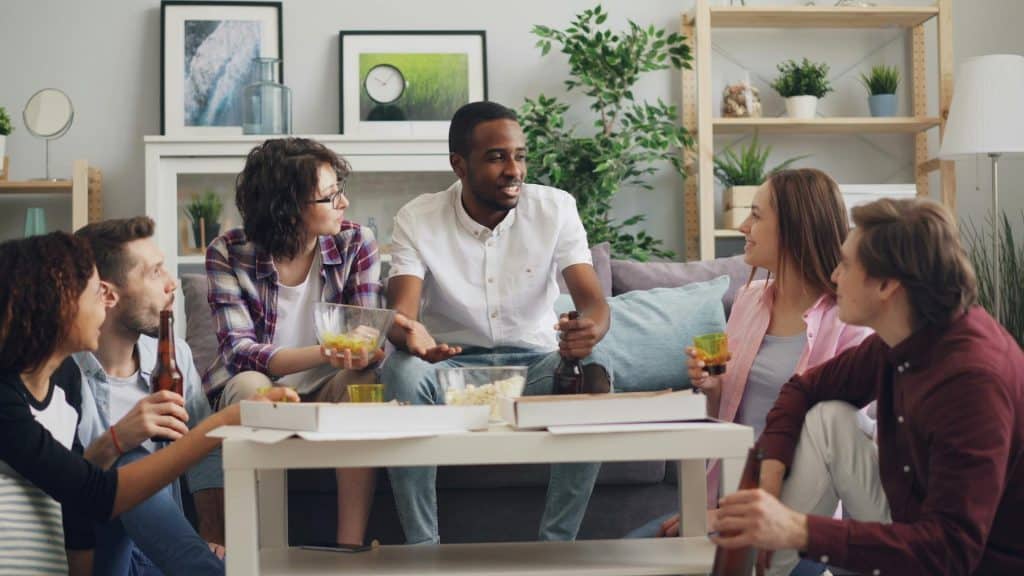 The group of people are gathered in the living room sharing drinks and food.