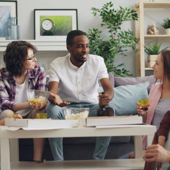 The group of people are gathered in the living room sharing drinks and food.
