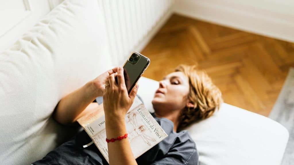 A person lying on a couch looking at a phone with a book resting on their chest.