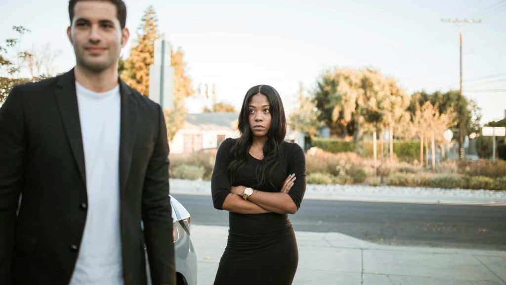 A woman standing with her arms crossed while a man walks away outdoors.